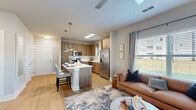 This thoughtfully designed 1-bedroom, 1-bathroom residence spans a single floor and maximizes every square foot with a smart, open-concept layout. The kitchen is a standout feature, showcasing warm wood-toned cabinetry, a full suite of stainless steel appliances including a French door refrigerator, electric range, microwave, and dishwasher, and a generous island with a white quartz countertop that doubles as a breakfast bar with seating. Stylish pendant lighting illuminates the space, which flows seamlessly into the living room. The living area benefits from large windows dressed with blinds and floor-length curtains, filling the space with natural light and offering outdoor views. Light wood-style flooring extends throughout the main living areas, adding warmth and continuity. The bedroom is a comfortable retreat featuring plush carpeting, a ceiling fan, and a window with outdoor views. A dedicated closet provides organized storage. The bathroom is clean and functional, featuring a wood-toned vanity with a white countertop, an undermount sink, and a full tub-shower combination. The overall ambiance is modern and inviting, characterized by a neutral color palette, contemporary finishes, and a cohesive design that balances style with practicality.