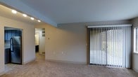 This single-floor space features a straightforward, open layout designed for functional living. The main living area is finished with wall-to-wall carpeting in a neutral tone, contributing to a quiet, comfortable atmosphere. Smooth, light-painted walls and a clean white baseboard trim give the space a simple, understated aesthetic.

A sliding glass door fitted with vertical blinds occupies one wall, allowing natural light to filter into the room while offering access to an outdoor area. The door provides a view of surrounding greenery, adding a subtle connection to the outdoors.

Recessed track lighting is installed in a lowered ceiling section near the entryway to an adjoining room, which appears to serve as a bathroom or utility space. That secondary area includes a window that introduces additional natural light, a glimpse of cabinetry, and what appears to be a sink, suggesting practical functionality.

The overall ambiance of the space is clean and neutral, with minimal ornamentation. The consistent color palette of soft grays and beiges throughout the walls, ceiling, and flooring creates a cohesive, move-in-ready environment suited for a variety of furnishing styles.