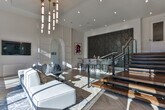 Located at 6000 Summit Place in Alpharetta, Georgia, this elegantly designed clubhouse showcases a seamless blend of modern luxury and refined architectural detail. The space is anchored by soaring double-height ceilings that amplify the sense of openness, while expansive floor-to-ceiling windows flood the interior with natural light and offer sweeping views of the surrounding landscape. Distinctive light herringbone-pattern hardwood flooring runs throughout, adding warmth and visual texture to the bright, airy environment. The main lounge area is appointed with contemporary sectional seating beneath a striking multi-tiered brass and glass pendant chandelier, creating a welcoming yet upscale gathering space. A dramatic staircase, framed by glass panel railings and rich wood treads, is flanked by two bold accent walls — one featuring large-scale veined marble-look tile, and another adorned with an artistic honeycomb-patterned wood panel, lending an artful, gallery-like quality to the space. A bar area with tall upholstered seating provides an inviting spot for socializing. The entryway is equally impressive, with its soaring clearstory windows, crisp white walls, and views into an adjacent fitness area. Elegant arched architectural details and curated artwork complete the polished, sophisticated ambiance throughout.