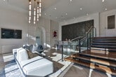 Located at 6000 Summit Place in Alpharetta, Georgia, this elegantly designed clubhouse showcases a seamless blend of modern luxury and refined architectural detail. The space is anchored by soaring double-height ceilings that amplify the sense of openness, while expansive floor-to-ceiling windows flood the interior with natural light and offer sweeping views of the surrounding landscape. Distinctive light herringbone-pattern hardwood flooring runs throughout, adding warmth and visual texture to the bright, airy environment. The main lounge area is appointed with contemporary sectional seating beneath a striking multi-tiered brass and glass pendant chandelier, creating a welcoming yet upscale gathering space. A dramatic staircase, framed by glass panel railings and rich wood treads, is flanked by two bold accent walls — one featuring large-scale veined marble-look tile, and another adorned with an artistic honeycomb-patterned wood panel, lending an artful, gallery-like quality to the space. A bar area with tall upholstered seating provides an inviting spot for socializing. The entryway is equally impressive, with its soaring clearstory windows, crisp white walls, and views into an adjacent fitness area. Elegant arched architectural details and curated artwork complete the polished, sophisticated ambiance throughout.
