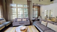 Located at 978 Westcreek Lane in Thousand Oaks, California, this sophisticated single-story space makes a strong first impression with a welcoming entryway featuring dark hardwood floors, soft gray walls, transom windows that draw in natural light, and a neatly appointed console area flanked by framed artwork.

The main living area is defined by dramatic soaring ceilings with rich, dark exposed wood beams that add architectural character and warmth. An expansive wall of multi-pane, grid-style windows spans the length of the room, framing views of mature trees and lush greenery while flooding the interior with abundant natural light. Elegant floor-length drapes in warm taupe frame the windows and contribute to the refined ambiance.

Dark hardwood flooring flows continuously throughout, grounding the open layout. The living space is styled with plush gray upholstered seating, a striped yellow-and-gray area rug, and a sculptural round coffee table, creating an inviting yet polished atmosphere. A media console with a wall-mounted flat-screen TV anchors one wall, while a quiet reading nook with a classic accent chair and curated artwork offers a more intimate retreat within the space.

The open floor plan connects seamlessly to a dining area and kitchen beyond, making this a highly functional and visually cohesive environment throughout.