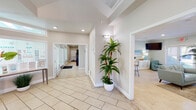 This single-story community clubhouse is designed with a coastal aesthetic that is both inviting and functional. The entry foyer sets an elegant tone with soaring ceilings, large-format light tile flooring laid in a diagonal pattern, a statement pendant light, and tall potted tropical plants flanking the passage into the main lounge area. Natural light filters in through transom windows, enhancing the bright, airy atmosphere throughout.

The lounge is a standout gathering space, featuring light hardwood flooring, soft seafoam-blue leather sofas, and a striking herringbone-tiled fireplace as the focal point. A wall-mounted television and access to outdoor views complete the relaxed, resort-like ambiance.

The leasing office area offers a clean, professional environment with large windows, light tile flooring, a welcoming reception desk, and ample natural light. Adjacent to this is a compact, well-appointed kitchen featuring white cabinetry, stainless steel appliances, stone countertops, and a pass-through breakfast bar.

The fitness center is divided into two well-equipped zones: one housing cardio machines including treadmills, stair climbers, and rowers with wall-mounted TVs and mirrored walls, and the other featuring a multi-use cable machine, free weights, and medicine balls. Large windows bring in natural light and outdoor views, while ceiling fans maintain comfort during workouts.