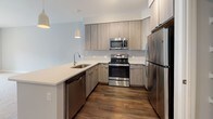 Located at 2614 Smooth Alder St in Gambrills, Maryland, this single-floor residence offers a clean, contemporary design throughout. The open-concept layout connects the living area to a well-appointed kitchen, creating a spacious and functional flow. The kitchen features light wood cabinetry extending to the ceiling, sleek white quartz countertops, stainless steel appliances including a range, microwave, dishwasher, and refrigerator, and rich wood-look flooring underfoot. Pendant lighting adds a modern touch above the kitchen peninsula. The living area is finished with plush neutral carpeting and soft gray walls that lend a calm, cohesive ambiance. Sliding glass doors lead to a private balcony overlooking a community pool and surrounding buildings, inviting ample natural light into the space. The bathroom is outfitted with a light wood vanity, white quartz countertop, an undermount sink with contemporary chrome fixtures, a large mirror, and neutral tile flooring. The overall design palette — warm wood tones, soft grays, and crisp white surfaces — creates a modern and inviting atmosphere throughout this well-designed Gambrills residence.
