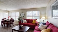 This single-floor residence offers a comfortable and functional layout with distinct living, dining, kitchen, and sleeping areas. The living room serves as the heart of the home, featuring durable wood-style flooring, multiple windows with horizontal blinds that invite generous natural light, and a textured ceiling. The space is generously sized to accommodate ample seating arrangements. Adjacent to the living area, the dining space benefits from large sliding glass doors dressed with vertical blinds alongside a window, providing an open view of the surrounding greenery outside. The kitchen is appointed with warm wood-toned cabinetry, white countertops, a double-basin sink, and full-size appliances including a refrigerator and range, all set above matching wood-style flooring. The primary bedroom features plush carpeting, a ceiling fan with light fixture, and direct access to a vanity area, offering added convenience. A second bedroom, also carpeted and equipped with a ceiling fan, provides a cozy and versatile space. A hallway connects the bedrooms to the bathroom, which includes a shower. Throughout the residence, neutral wall tones create a cohesive and welcoming ambiance.