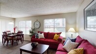 This single-floor residence offers a comfortable and functional layout with distinct living, dining, kitchen, and sleeping areas. The living room serves as the heart of the home, featuring durable wood-style flooring, multiple windows with horizontal blinds that invite generous natural light, and a textured ceiling. The space is generously sized to accommodate ample seating arrangements. Adjacent to the living area, the dining space benefits from large sliding glass doors dressed with vertical blinds alongside a window, providing an open view of the surrounding greenery outside. The kitchen is appointed with warm wood-toned cabinetry, white countertops, a double-basin sink, and full-size appliances including a refrigerator and range, all set above matching wood-style flooring. The primary bedroom features plush carpeting, a ceiling fan with light fixture, and direct access to a vanity area, offering added convenience. A second bedroom, also carpeted and equipped with a ceiling fan, provides a cozy and versatile space. A hallway connects the bedrooms to the bathroom, which includes a shower. Throughout the residence, neutral wall tones create a cohesive and welcoming ambiance.