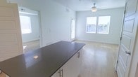 This single-floor space showcases a clean, contemporary design with an open-concept layout that seamlessly connects the kitchen and living areas. The kitchen features a substantial island with a dark countertop and crisp white cabinetry fitted with sleek bar-pull hardware, providing both prep space and a natural visual anchor for the room. Light hardwood flooring extends throughout the main living area, contributing to the space's airy, cohesive feel.

Large windows dressed with white horizontal blinds allow abundant natural light to fill the living area, enhancing the bright, white wall palette. A ceiling fan with a light fixture is centrally positioned, offering both functional ventilation and subtle illumination. A glass-paneled door provides additional light and likely leads to an outdoor area.

An adjacent room, partially visible through a doorway, features carpeted flooring, offering a softer, quieter environment suitable for a bedroom or den. The overall ambiance is modern and minimalist, with a neutral color scheme that creates a versatile, move-in-ready environment adaptable to a variety of interior styles.