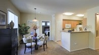 This single-floor residence showcases an open-concept design that seamlessly connects the main living areas. The dining area is anchored by a round glass-top table surrounded by upholstered patterned chairs, lit by a warm pendant light fixture overhead. A decorative floral arrangement and large tropical houseplant add natural vibrancy to the space.

Adjacent to the dining area, the kitchen features light maple cabinetry, white appliances including a microwave and range, and a raised breakfast bar that serves as a natural divider between the cooking and dining spaces. The countertops provide ample workspace, and the layout promotes easy interaction between areas.

The open sightline from the dining space leads directly into a bedroom, visible through a doorway and furnished with a bed and window treatments that allow controlled natural light. An additional door nearby suggests further storage or utility access.

Throughout, neutral-toned carpeting and light-colored walls create a cohesive, understated aesthetic. The layout is practical and well-organized, offering a comfortable flow between functional living spaces on a single level.