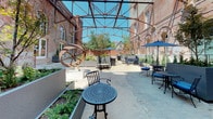 The Brewery Courtyard is a thoughtfully restored open-air space that seamlessly blends industrial heritage with contemporary outdoor amenities. Framed by the original multi-story red brick building facade, the courtyard retains much of its historic character, highlighted by arched windows, original masonry, and an iconic overhead steel truss framework that defines the space while remaining open to the sky above.

The ground level features a combination of classic cobblestone and smooth concrete paving, creating distinct zones for gathering and relaxation. Black wrought iron bistro tables and chairs, accented with blue cushions and complementing patio umbrellas, are arranged throughout to encourage both intimate conversation and larger group settings. Lounge seating areas provide additional comfort for more casual use.

Generous tiered concrete planters are a defining landscape element, showcasing ornamental trees, shrubs, and ground-level ferns that bring lush greenery to the hardscape environment. A standout sculptural metal kinetic art piece adds a distinctive artistic focal point to the courtyard.

The space benefits from abundant natural sunlight throughout the day, while the surrounding brick walls create a sense of enclosure and shelter. A railed balcony-level walkway connects the courtyard to the adjacent building interiors, reinforcing the space's role as a vibrant, accessible hub within the larger brewery complex.