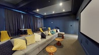 This single-floor screening room is thoughtfully designed to deliver a true cinema-quality experience within a private setting. The space is anchored by a large, wall-mounted projection screen positioned at one end of the room, flanked by dark-painted walls that enhance contrast and minimize light interference during viewing. Recessed ceiling lights, set within a coffered ceiling detail, provide adjustable ambient illumination, while a subtle blue LED strip traces the upper perimeter of the room, adding a stylish atmospheric glow.

The primary seating arrangement consists of a generously sized, light gray sectional sofa positioned centrally and angled toward the screen. Colorful throw pillows in yellow, teal, navy, and white accent the neutral upholstery, adding visual warmth. Compact, round wooden side tables are scattered throughout for convenience. Behind the main sectional, a secondary lounge zone features freestanding chairs draped with yellow throws, alongside additional small tables for casual use.

Along the back wall, floor-to-ceiling charcoal curtains frame what appears to be windows or a decorative drapery treatment, contributing to the room's light-controlled, theatrical ambiance. Small bistro-style tables with traditional chairs occupy this rear zone, offering an alternative seating option. The carpeted flooring throughout absorbs sound while enhancing the overall acoustic quality of this well-appointed screening space.