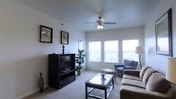 This single-floor space centers around a comfortable living room designed with both functionality and a relaxed ambiance in mind. The room features neutral-toned carpet flooring and smooth, light-colored walls that create a clean, airy atmosphere. A ceiling fan with an integrated light fixture is mounted overhead, providing both air circulation and supplemental lighting. Large windows along one wall allow generous amounts of natural light to fill the room, brightening the space and contributing to its open feel. Horizontal blinds on the windows offer privacy and light control. The living area is furnished with a plush sectional sofa arrangement and a glass-topped coffee table, creating a practical and inviting seating area. A dark-finished media console houses a flat-screen television, serving as a natural focal point opposite the seating. Decorative touches include framed wall art, a tall decorative vase, and an indoor potted plant that adds a subtle natural element to the space. The overall design aesthetic is understated and functional, with a neutral color palette that provides a versatile backdrop for various décor styles.