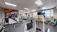 This fitness center offers a comprehensive workout environment organized into distinct functional zones across a single floor. The cardio area features a row of treadmills and elliptical trainers, each equipped with individual screens for entertainment during exercise. A staircase nearby provides an additional access point and adds architectural interest to the space.

The strength training area is well-appointed with a multi-station cable machine, a Smith machine loaded with Olympic plates, and a full rack of dumbbells ranging across various weight increments. Flat and adjustable benches are positioned throughout for versatile use. A heavy punching bag on a freestanding mount adds a boxing component to the training options.

A dedicated functional training corner houses stability balls, resistance bands, foam rollers, kettlebells, and other accessories organized neatly on racks. Wall-mounted flat-screen televisions are placed strategically throughout the space.

Large mirrors line key walls, visually expanding the room and allowing users to monitor their form. Recessed fluorescent panel lighting provides consistent, even illumination throughout. A standout visual feature is the bold, colorful "Charm City" graffiti-style mural, adding a vibrant, locally inspired identity to the space. Rubber flooring covers the entire area, ensuring safety and durability.