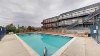 Located at 13050 W Cedar Dr in Lakewood, Colorado, this outdoor amenity area centers around a generously sized swimming pool with vivid turquoise water, offering a refreshing retreat for residents. The pool is rectangular in form and framed by a broad concrete deck that provides ample space for lounging and socializing.

The sundeck is well-furnished with numerous sling-style lounge chairs arranged along the poolside, ideal for relaxing in the Colorado sun. Several patio tables with chairs are also positioned around the deck, offering comfortable spots for outdoor dining or casual gatherings.

The entire pool area is enclosed by a sturdy black metal fence, ensuring both security and safety. Entry points are gated, maintaining a controlled and private environment. The surrounding landscape includes mature trees and open greenery, contributing to a pleasant, park-like atmosphere.

The multi-story residential buildings adjacent to the pool feature wood-railed balconies and staircases that overlook the amenity area, creating a sense of community connectivity. The architectural style of the buildings reflects a classic mid-century design with brick and wood detailing beneath dark shingled rooflines. Open skies above offer views of the broader Lakewood landscape, making this pool area a standout outdoor feature of the property.