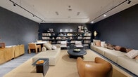 Located at 2140 N Miami Ave in Miami's vibrant Wynwood neighborhood, this single-floor space delivers a refined, lounge-like atmosphere with a distinctive aesthetic. The interior is defined by deep charcoal and dark gray walls that create a moody, intimate ambiance, contrasted by light gray hardwood flooring and a neutral area rug that anchors the central seating arrangement. Track lighting runs across the white ceiling, casting focused illumination throughout the room and highlighting key design elements.

The seating area serves as the heart of the space, featuring a large cream modular sofa paired with a cognac leather accent chair and a sculptural round black coffee table. Two mid-century-style armchairs with striped upholstery add additional seating near the back. A warm wood credenza with arched panel detailing lines one wall, topped with a vintage record player, decorative objects, and florals.

Floor-to-ceiling open metal shelving dominates the rear wall, displaying an extensive vinyl record collection alongside curated books, framed artwork, and decorative accents — reinforcing the space's music-centric identity. A compact bistro table with chairs offers an intimate seating nook. The overall design balances curated eclecticism with a cool, editorial sensibility suited to its dynamic Miami location.
