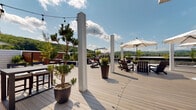 Located at 400 Boulder Pass in Oxford, Connecticut, this remarkable outdoor space is a generously sized rooftop deck designed for relaxed gatherings and al fresco living. The deck is finished with wide-plank composite decking in a warm gray tone, providing a clean, contemporary foundation throughout the space.

Elegant white structural columns are positioned across the deck, strung with bistro-style string lights that lend a welcoming ambiance for evening use. Multiple large market umbrellas offer shaded seating areas, complemented by Adirondack chairs, lounge chairs, and conversation sets arranged to take full advantage of the open-air setting. Potted ornamental trees and lush plantings add a natural, landscaped feel to the rooftop environment.

A standout feature is the striking natural stone chimney rising alongside blue-gray siding, anchoring one corner of the space with architectural character. A ping-pong table provides a recreational focal point, while a dedicated dining area with bar-height seating accommodates entertaining. The deck wraps around to include a hot tub area framed by warm wood privacy panels.

Panoramic views of the rolling, tree-covered Connecticut hills stretch out in every direction, making this rooftop deck an exceptional retreat that seamlessly blends sophisticated outdoor design with the natural beauty of its surroundings.