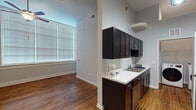 This single-floor unit combines functional design with practical finishes throughout its well-organized layout. The galley-style kitchen is a standout feature, offering dark espresso cabinetry lining both sides, white countertops, and a full suite of stainless steel appliances including a refrigerator, electric range, and built-in microwave. A stainless steel sink and dishwasher add to the kitchen's utility, while in-unit front-loading washer and dryer are conveniently located at the rear of the kitchen corridor. Warm hardwood flooring runs continuously through the kitchen and living areas, contributing to a cohesive, inviting atmosphere. The living area is generously proportioned, bathed in natural light from large windows fitted with horizontal blinds, and topped with a ceiling fan for year-round comfort. The bathroom is well-appointed with dark cabinetry beneath a white countertop vanity, a large mirror, a tub-shower combination, and distinctive patterned tile flooring. A dedicated walk-in closet provides substantial storage, outfitted with wire shelving and carpeted flooring. Overall, the unit presents a clean, practical aesthetic suited for comfortable everyday living.