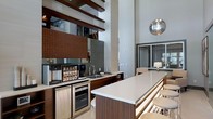 This single-floor space makes a strong architectural statement with its dramatically high ceilings that amplify the sense of openness and volume. The kitchen and bar area serves as a central focal point, featuring a long, clean-lined white countertop supported by rich dark wood cabinetry. The design is further elevated by warm-toned mosaic tile backsplashes in deep brown hues, adding texture and visual depth to the space.

The kitchen area is well-equipped, with built-in appliances including a beverage station, compact refrigeration, and an oven integrated seamlessly beneath the counter. A stainless steel sink is neatly positioned along the workspace, maintaining the space's clean, modern aesthetic. Bar-height seating lines the counter, making it ideal for casual dining or socializing.

A striking chrome spherical pendant light serves as a bold decorative accent, catching light and contributing to the contemporary atmosphere. Exposed horizontal beams near the ceiling add an industrial design element that complements the modern finishes. In the background, a framed opening reveals an adjacent lounge area with warm lamp lighting, hinting at the space's versatility and cohesive design throughout.