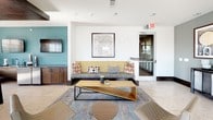 Located at 305 W Commerce St in Dallas, TX, this single-floor community lounge offers a well-appointed gathering space designed for both relaxation and social interaction. The interior features a cohesive contemporary aesthetic, anchored by large-format tile flooring, warm wood accents, and a sophisticated palette of slate gray, teal, and crisp white throughout.

The lounge is divided into distinct yet open zones that flow seamlessly into one another. A casual seating area centers around a low-profile wooden coffee table and a plush sofa with colorful accent pillows, positioned beneath a wall-mounted flat-screen TV. A Dallas city map serves as local artwork, reinforcing the space's connection to its surroundings. Natural light pours in through generous windows that frame views of the Dallas skyline.

An entertainment zone features a shuffleboard table, additional lounge seating with patterned throw pillows, and curated framed artwork along the walls, adding a gallery-like quality to the space. Pendant lighting with warm-toned drum shades and a statement sputnik chandelier provide ambient illumination, complemented by recessed wood ceiling insets that add architectural depth.

A dining area with a solid wood table, mid-century modern chairs, and a marble-topped bar counter offers a functional space for dining or casual meetings. A compact service counter with a mini-fridge rounds out the amenities, making this lounge a fully equipped and inviting community hub.