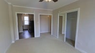 This single-floor space is designed with a practical, open layout that connects its key living areas seamlessly. The main room features wall-to-wall carpeting in a neutral beige tone, complemented by warm taupe walls and clean white trim that frame each doorway and architectural detail. Crown molding runs along the ceiling, adding a subtle classic touch, while a ceiling fan with a central light fixture provides both ventilation and ambient lighting.

A set of mirrored sliding closet doors lines one wall of the main room, offering generous built-in storage while visually expanding the sense of space. An open doorway leads to an adjacent room — potentially a second bedroom or flexible-use space — also carpeted and finished in matching neutral tones with a pendant light.

The kitchen, accessible through an arched doorway, transitions to dark tile flooring and features black appliances, including a full-size refrigerator. A window with arched detailing allows natural light to fill the kitchen. The overall ambiance of the space is clean, understated, and move-in ready, with consistent finishes throughout that create a cohesive, functional living environment.