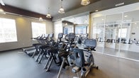 This fitness center is thoughtfully designed to support a comprehensive workout experience. The main cardio area is furnished with a row of Cybex machines, including treadmills, ellipticals, and stationary bikes, all arranged to face large flat-screen televisions mounted on the wall, making workouts more engaging. The space features a neutral color palette with dark accent walls and a dark ceiling that creates a sleek, modern atmosphere. Decorative pendant lights hang from the ceiling, providing warm, focused illumination that complements the recessed lighting throughout. Large windows along one wall allow natural light to fill the space, contributing to an open and inviting feel. Full-length mirrors line the walls, visually expanding the room and allowing users to monitor their form during exercise. A glass-paneled door leads to an adjoining room, visible in the background, which appears to offer additional fitness or studio space. The flooring is finished with durable, cushioned gym-grade material appropriate for high-traffic use. Overall, the space balances functionality with a polished, upscale aesthetic.