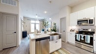 This thoughtfully designed one-bedroom, one-bathroom unit in Georgetown, Texas offers a bright, open-concept layout with modern finishes throughout. Light wood-tone flooring flows consistently across the living spaces, complementing the neutral color palette and creating a cohesive aesthetic.

The kitchen is a standout feature, showcasing white shaker cabinetry extending to the ceiling for generous storage, a herringbone tile backsplash, quartz countertops, and a full suite of stainless steel appliances including a refrigerator with ice maker, range, microwave, and dishwasher. A central island with an apron-front farmhouse sink and pendant lighting above serves as both a functional workspace and a casual dining area.

The open living area benefits from large windows and glass doors that flood the space with natural light and offer views of the surrounding neighborhood. The bedroom is a comfortable retreat with dual windows, wood-look flooring, and ample room for furnishings.

The bathroom features a long white vanity with dual undermount sinks, a large mirror, and wood-look flooring. Directly accessible from the bathroom is a well-organized walk-in closet with built-in shelving, hanging rods, and corner shelving units, providing excellent storage capacity.
