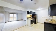 This single-floor residence features an open-concept design that seamlessly connects the kitchen and living area, creating a cohesive and functional living space. The kitchen is equipped with dark upper cabinetry, a stainless steel refrigerator, an electric range with an overhead ventilation hood, and light-toned vinyl flooring. Dark cabinet finishes provide a striking contrast against the white walls and light-colored surfaces. Adjacent to the kitchen, the living area is carpeted and benefits from natural light streaming through sliding glass doors that open onto an outdoor deck, offering views of the surrounding landscape. The ceiling features a textured finish throughout, complemented by a ceiling fan in the living space for added comfort. Clean white walls, practical baseboards, and straightforward lighting fixtures contribute to the space's understated and utilitarian aesthetic. The transition between the kitchen's vinyl flooring and the living area's carpet is clearly defined, lending each zone its own distinct character while maintaining an open and connected feel overall.