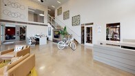 Located at 530 11th Ave N in Nashville's vibrant Gulch neighborhood, this contemporary single-floor commercial lobby combines industrial design with modern sophistication. Polished concrete floors extend throughout the entire space, complementing the soaring double-height ceilings that flood the interior with natural light through expansive floor-to-ceiling glass windows and doors. The entry vestibule welcomes visitors with bold abstract artwork, concrete columns, and direct views of the surrounding urban streetscape. A central open-plan lounge area is furnished with warm tan leather seating arrangements set atop golden-yellow area rugs, creating an inviting gathering space bathed in natural light from the oversized windows overlooking 11th Avenue. A striking wood-and-glass barn-style sliding door divides the main floor, adding an architectural focal point alongside a walnut-topped bar counter with seating — ideal for casual meetings or collaborative work. An upper mezzanine level with glass railings overlooks the main floor, enhancing the sense of vertical space. A dedicated coffee bar, outfitted with a Lavazza machine, dark wood cabinetry, and wall-mounted organizer racks, provides a functional refreshment nook. Curated artwork and decorative accents throughout contribute to a polished yet approachable ambiance.