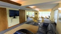 This single-floor space is thoughtfully designed to serve as a multifunctional lounge and gathering area. The layout is divided into distinct zones, each serving a specific purpose while maintaining a cohesive and welcoming atmosphere throughout.

At the forefront of the space is a relaxed lounge area anchored by a dark leather sofa and a patterned area rug, providing a comfortable setting for casual conversation or relaxation. A large flat-screen television is mounted on the wall nearby, complemented by a wooden media cabinet, making this corner ideal for entertainment.

Deeper into the space, a dining and social zone features multiple round tables surrounded by modern yellow chairs with chrome legs, offering ample seating for group gatherings or meetings. The furniture arrangement encourages interaction and flexibility in how the space is used.

Large windows line the far wall and wrap around the perimeter, flooding the interior with abundant natural light and offering views of the surrounding greenery outdoors. Exposed wooden ceiling beams add a warm, architectural character to the space, contrasting nicely with the light yellow walls and carpeted flooring. The overall ambiance is bright, open, and functional — well-suited for community events, meetings, or everyday social use.