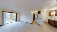 This single-floor space offers a straightforward and functional layout designed for comfortable everyday living. The main living area is finished with neutral wall-to-wall carpeting that creates a warm, inviting atmosphere, while large sliding glass doors along one wall flood the space with natural light and provide a view of the surrounding greenery. The open sightlines between the living area and kitchen promote an easy flow throughout the home. The kitchen features tile flooring and is equipped with white appliances, including a refrigerator, set against cabinetry finished in a rich, dark wood tone. A hallway visible from the main living area connects to additional rooms, with dark-trimmed doors adding a subtle contrast to the light-colored walls throughout. The textured ceiling and neutral color palette lend a clean, understated aesthetic to the interior. Floor vents are positioned throughout, and recessed lighting provides consistent illumination across the space. Overall, the unit presents a practical and well-organized floor plan that balances openness with defined functional zones, making it a versatile space suitable for a variety of living arrangements.