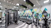 This fitness center offers a thoughtfully designed workout space that combines functionality with contemporary aesthetics. The room features a striking geometric mural along the back wall, rendered in teal, yellow, and white triangle patterns, adding a bold visual energy to the environment. Mirrored walls line one side of the space, visually expanding the room while allowing users to monitor their form during exercise.

The cardio section is well-equipped with a row of modern treadmills and an elliptical trainer, each outfitted with digital display consoles. A multi-functional cable machine and additional strength training equipment are positioned along the mirrored wall, providing a range of resistance training options.

Overhead, a suspended black-framed structure adorned with lush trailing greenery serves as a distinctive design element, softening the space with natural texture. String-style pendant lights and track lighting illuminate the room with a warm yet bright glow. The ceiling incorporates a drop-tile grid with integrated skylights, introducing natural light from above. The flooring features a striped grey pattern, contributing to the clean, modern finish of the overall space.