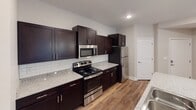 Located in Fort Mill, SC, this single-floor home offers a well-designed layout with thoughtful finishes throughout. The kitchen is a standout feature, boasting rich dark espresso cabinetry, granite-look countertops, a classic white subway tile backsplash, and a full suite of stainless steel appliances including a range, microwave, and refrigerator. A peninsula with an integrated sink adds both functionality and additional workspace. Wood-look plank flooring runs through the main living and kitchen areas, lending warmth and continuity to the open layout. The living area is bright and open, with natural light filtering through large windows. Bedrooms are carpeted and feature ceiling fans with light fixtures and windows overlooking greenery outside. A generously sized walk-in closet with wire shelving and basket storage provides ample organization space. The primary bathroom is well-appointed with dark cabinetry, a wide vanity with a large mirror, a three-bulb vanity light fixture, and a separate glass-enclosed shower. A secondary bathroom offers a full tub-shower combo, dark vanity, and tile flooring. Completing the home is a dedicated laundry room with a full-size top-loading washer and dryer and overhead wire shelving for added storage.