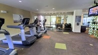 This fitness center offers a thoughtfully arranged workout space designed to accommodate a variety of exercise routines. The room features a selection of modern cardio equipment, including treadmills and elliptical machines, positioned near large windows that allow natural light to fill the space and provide views of the surrounding outdoors. The flooring consists of durable, patterned carpet with colorful accent squares in orange, yellow, and green, adding a dynamic visual element to the room.

Along one wall, floor-to-ceiling mirrors create a sense of expanded space while allowing users to monitor their form during exercise. A well-stocked free weight area lines the mirrored wall, featuring multiple dumbbell racks with a wide range of weights to suit various fitness levels. Recessed lighting and track lighting are installed throughout the ceiling, ensuring consistent, even illumination across the entire floor.

The neutral-toned walls and clean layout contribute to a functional and focused atmosphere. The space is designed to serve as a comprehensive fitness facility, balancing cardiovascular and strength training equipment in an organized, accessible environment.