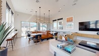 This well-appointed single-floor community space offers a thoughtfully designed open-concept layout that seamlessly blends lounge, dining, and kitchen functions. Soaring ceilings define the main gathering area, amplifying the sense of spaciousness throughout. The lounge zone features a mix of plush sectional sofas, rattan accent chairs, and a large flat-screen TV mounted above a sleek media console, complemented by a built-in wooden shelving unit displaying curated décor. A large ceiling fan overhead adds both style and comfort.

The dining area is a standout feature, anchored by a curved cognac leather banquette seating arrangement paired with white marble-topped tables and contemporary dark chairs. Striking sputnik-style pendant chandeliers hang above, adding a bold design focal point. Floor-to-ceiling sliding glass doors and expansive windows flood the space with natural light and provide direct views and access to an outdoor patio area with additional seating and umbrellas.

The kitchen is finished with crisp white cabinetry, a sage green geometric tile backsplash, stainless steel appliances including a French door refrigerator, and open wooden floating shelves. Light-toned large-format tile flooring runs throughout the entire space, tying all zones together cohesively. Black-framed glass doors add a modern industrial touch at key transitions throughout the space.