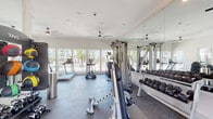This single-floor fitness center is thoughtfully designed to accommodate a comprehensive range of exercise needs. The space is bright and inviting, with large windows lining the walls and allowing abundant natural light to flood the interior while offering views of the surrounding outdoor community. Ceiling fans are installed throughout, promoting airflow and comfort during workouts.

The cardio zone is equipped with multiple treadmills and elliptical machines from True Fitness, positioned to take advantage of the natural light and outdoor views. A multi-functional cable machine serves as the centerpiece for strength training, complemented by a full rack of dumbbells in graduated weights neatly arranged along a mirrored wall. The floor-to-ceiling mirrors visually expand the space and allow users to monitor their form. Additional equipment includes a Hoist machine, weight bench, and a medicine ball and stability ball rack offering functional fitness options.

The flooring throughout is durable rubber gym flooring, providing traction and impact absorption. The neutral-toned walls and recessed lighting contribute to a clean, modern aesthetic. Entry to the fitness center is accessible via glass-panel doors that connect directly to the outdoor grounds, further enhancing the open, airy feel of the space.