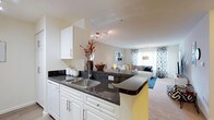 This single-floor residence offers a thoughtfully designed open-concept layout that seamlessly connects the kitchen and living areas, creating a cohesive and functional living environment. The kitchen is outfitted with crisp white cabinetry, dark granite countertops, and a stainless steel sink, all centered around a peninsula that doubles as a breakfast bar — an ideal spot for casual dining or entertaining. Track lighting above the kitchen adds a modern touch while ensuring excellent task illumination. The living area extends naturally from the kitchen, featuring warm neutral wall tones and plush carpeting that contribute to a comfortable, inviting ambiance. Large windows dressed with patterned curtains allow generous natural light to fill the space, enhancing its open and airy feel. The overall design strikes a balance between contemporary style and practical functionality, with clean lines and a neutral palette that lends itself well to a variety of décor preferences. The flow between the kitchen and living room makes the space feel expansive and well-suited for both relaxed daily living and hosting guests.
