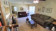This single-floor residence offers a cozy and well-appointed living environment designed for comfort and everyday functionality. The living room serves as the heart of the space, featuring plush wall-to-wall carpeting that adds warmth underfoot. Two ceiling light fixtures — a chandelier pendant and a ceiling fan with integrated lighting — provide ample illumination throughout the room, creating a welcoming ambiance at any time of day. The room is generously sized, comfortably accommodating a full sofa and a loveseat arranged around a dark wood coffee table, making it well-suited for both relaxing and entertaining. A sliding glass door allows natural light to filter into the space and offers a direct connection to the outdoors. Framed artwork adorns the warm-toned walls, adding a personal and lived-in character to the interior. A doorway visible from the living area suggests additional rooms beyond, hinting at a thoughtfully laid-out floor plan. The neutral color palette of the walls and carpet creates a versatile backdrop that ties the space together cohesively. Overall, this residence presents a comfortable, functional, and homey atmosphere throughout its single level.