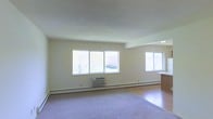 This single-floor space features an open-concept living area designed to maximize natural light and functionality. The main living space is generously proportioned, with large windows along the exterior wall that invite ample daylight and offer pleasant views of mature trees and green surroundings outside.

The living area is finished with wall-to-wall carpeting, providing a comfortable and warm underfoot feel, while an adjacent area transitions to hardwood-style flooring, adding visual contrast and variety to the overall aesthetic. The neutral, cream-toned walls keep the space feeling clean, bright, and versatile, accommodating a wide range of furnishing styles.

Climate control is well-addressed with a wall-mounted air conditioning unit positioned beneath the main window grouping, supplemented by baseboard heating that runs along the perimeter of the room, ensuring year-round comfort. A counter or breakfast bar is partially visible toward the right side of the space, suggesting a kitchen or dining area just beyond the main living room. The textured ceiling adds subtle architectural detail, while overall, the space maintains a straightforward, functional layout well-suited for everyday living.