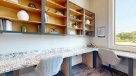 This business center office is a thoughtfully designed workspace that maximizes functionality within a compact footprint. The main work area features a continuous L-shaped countertop finished in speckled granite, supported by teal-accented cabinetry that adds a modern, polished aesthetic. Built-in open shelving units with warm wood interiors and teal frames line the wall above the workstation, providing ample display and storage space for books, decor, and office essentials. Ergonomic task chairs are positioned throughout, accommodating multiple users simultaneously. A large window at the corner of the workstation floods the space with natural light and offers a pleasant view of mature trees and greenery outside, creating an inviting atmosphere for focused work. The flooring consists of patterned carpet in muted gray tones, adding texture and warmth underfoot. Adjacent to the main workspace is a glass-panel conference room framed by rich, dark wood doors, offering a semi-private meeting area with seating for up to three occupants. The combination of natural light, quality materials, and purposeful layout makes this business center a highly functional and visually appealing space for professional use.