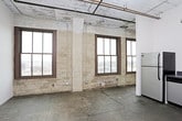 This single-floor space embraces an industrial loft aesthetic defined by raw, unfinished materials and a distinctly urban character. Polished concrete floors extend throughout, bearing the natural markings and texture of aged stone, while exposed masonry walls reinforce the building's historic origins. Visible ductwork and piping run along the ceilings, adding to the space's industrial authenticity.

The kitchen is fitted with a full suite of stainless steel appliances, including a range with oven, overhead microwave, refrigerator, and dishwasher. White flat-panel cabinetry lines the L-shaped layout, complemented by dark gray countertops that provide a clean contrast. Overhead fluorescent lighting illuminates the workspace efficiently.

The main living area is open and expansive, with soaring ceilings and large, grid-paned windows set within the exposed masonry walls. These windows draw in natural light and frame views of the surrounding urban landscape. The open floor plan allows for flexible furniture arrangements throughout the generous living space.

The bathroom corridor features light wood sliding doors and continues the concrete flooring found throughout the unit. An exposed structural column adds further architectural interest. A closet with built-in shelving is found in the bedroom, which also benefits from a large masonry-framed window with city views.