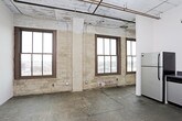 This single-floor space embraces an industrial loft aesthetic defined by raw, unfinished materials and a distinctly urban character. Polished concrete floors extend throughout, bearing the natural markings and texture of aged stone, while exposed masonry walls reinforce the building's historic origins. Visible ductwork and piping run along the ceilings, adding to the space's industrial authenticity.

The kitchen is fitted with a full suite of stainless steel appliances, including a range with oven, overhead microwave, refrigerator, and dishwasher. White flat-panel cabinetry lines the L-shaped layout, complemented by dark gray countertops that provide a clean contrast. Overhead fluorescent lighting illuminates the workspace efficiently.

The main living area is open and expansive, with soaring ceilings and large, grid-paned windows set within the exposed masonry walls. These windows draw in natural light and frame views of the surrounding urban landscape. The open floor plan allows for flexible furniture arrangements throughout the generous living space.

The bathroom corridor features light wood sliding doors and continues the concrete flooring found throughout the unit. An exposed structural column adds further architectural interest. A closet with built-in shelving is found in the bedroom, which also benefits from a large masonry-framed window with city views.