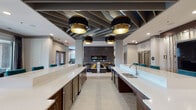 This single-floor amenity space is thoughtfully designed to accommodate both communal gatherings and quiet relaxation, with a layout that flows seamlessly between several distinct zones. The kitchen and entertaining area anchors the space with two elongated white quartz countertop islands, each flanked by teal upholstered barstools and equipped with stainless steel sinks and dishwashers. Dark walnut cabinetry provides warm contrast beneath the crisp counters, while a dramatic open ceiling with exposed steel beams and large black-and-gold drum pendant lights adds an industrial-chic character overhead. Large-format light tile flooring runs throughout, unifying the zones visually. A central lounge features built-in banquette seating with bold patterned upholstery, highlighted by yellow and teal accent pillows, surrounding a linear fireplace set within a full dark tile feature wall. Adjacent to this is a refined sitting area with a curved cream sectional, a large-scale floral artwork in black and gold, and sculptural décor pieces on a patterned area rug. A separate nook offers a dark bistro-height table with gold-toned stools beneath a wood ceiling accent panel, alongside a decorative wine rack display. A windowed seating bench near the entrance provides views of an outdoor pool area, filling the space with natural light.