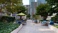 Located at 345 E Wacker Drive in the heart of Chicago, this outdoor patio offers a well-appointed urban retreat surrounded by the city's iconic skyline. The space is designed with large-format stone tile flooring that extends across a generous open-air terrace, providing ample room for both relaxed lounging and social gatherings. Thoughtfully arranged seating clusters feature modern aluminum chairs and tables in vibrant blue tones, accented by colorful market umbrellas in blue and chartreuse green that provide shade throughout the day. Raised concrete planters filled with lush ground cover, mature shade trees, and seasonal flowering plants define distinct zones within the patio, adding natural texture and a sense of greenery to the urban setting. A standout feature of the space is the built-in outdoor kitchen, equipped with multiple stainless steel gas grills set into wood-paneled cabinetry — ideal for communal cooking and entertaining. Additional lounge seating is tucked along the perimeter, offering quieter spots for relaxation. The patio borders the building's glass-clad facade, and a perimeter railing opens up views to the surrounding Streeterville neighborhood and Chicago's impressive high-rise skyline, creating a dynamic backdrop for this versatile outdoor amenity.