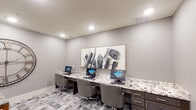 This well-appointed business center offers a polished and professional environment designed to support both focused individual work and collaborative meetings. The space is divided into two functional zones, seamlessly connected through a glass-paneled interior door that allows light to flow between areas.

The workstation area features a long, built-in desk with striking granite countertops and dark wood cabinetry below, providing ample storage. Multiple computer terminals are arranged side by side, each accompanied by ergonomic rolling chairs. A large, bold abstract artwork in black, white, and gold tones anchors the wall above the workstation, adding a sophisticated artistic touch. Recessed lighting overhead ensures a bright, well-lit workspace.

The adjacent meeting area accommodates small group gatherings around a marble-topped table paired with plush upholstered chairs. A dramatic oversized decorative clock with an open metal frame serves as a striking focal point on the wall. The glass-panel door entry leads to a connecting corridor, offering easy access between spaces.

Throughout, the consistent design language of soft gray walls, geometric patterned flooring in varying shades of gray, and clean white baseboards creates a cohesive, contemporary aesthetic. The overall ambiance is refined and functional, making it an ideal setting for productivity and professional interaction.