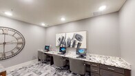 This well-appointed business center offers a polished and professional environment designed to support both focused individual work and collaborative meetings. The space is divided into two functional zones, seamlessly connected through a glass-paneled interior door that allows light to flow between areas.

The workstation area features a long, built-in desk with striking granite countertops and dark wood cabinetry below, providing ample storage. Multiple computer terminals are arranged side by side, each accompanied by ergonomic rolling chairs. A large, bold abstract artwork in black, white, and gold tones anchors the wall above the workstation, adding a sophisticated artistic touch. Recessed lighting overhead ensures a bright, well-lit workspace.

The adjacent meeting area accommodates small group gatherings around a marble-topped table paired with plush upholstered chairs. A dramatic oversized decorative clock with an open metal frame serves as a striking focal point on the wall. The glass-panel door entry leads to a connecting corridor, offering easy access between spaces.

Throughout, the consistent design language of soft gray walls, geometric patterned flooring in varying shades of gray, and clean white baseboards creates a cohesive, contemporary aesthetic. The overall ambiance is refined and functional, making it an ideal setting for productivity and professional interaction.