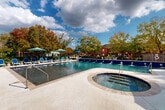 Located at 10720 Linkwood Ct in Baton Rouge, Louisiana, this outdoor amenity complex offers a resort-inspired experience centered around a generously sized swimming pool with elegant blue tile accents and built-in water features along its perimeter. Adjacent to the main pool sits a dedicated circular hot tub, complete with jets and easy-access handrails, providing a relaxing retreat for residents. The expansive pool deck is finished in light-toned pavers and surrounds the water on multiple sides, accommodating a large collection of blue lounge chairs and umbrella-shaded seating areas perfect for sunbathing and outdoor leisure. Mature trees and lush landscaping frame the perimeter, offering natural shade and a sense of privacy. The attached single-story clubhouse building showcases a classic brick exterior complemented by crisp white columns supporting a covered outdoor terrace. Large glass doors and windows allow natural light to flow freely, while the covered patio provides a comfortable transition between indoor and outdoor spaces. Additional outdoor seating, including Adirondack chairs and café-style tables, are thoughtfully arranged under the pavilion. The overall ambiance of this facility is bright, well-maintained, and inviting, making it a standout communal amenity within the Baton Rouge community it serves.