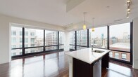 This contemporary single-floor residence offers a thoughtfully designed open-concept layout that seamlessly integrates the kitchen and living areas. Rich, dark hardwood flooring runs throughout the main living spaces, providing a warm contrast to the bright white ceilings and walls. Floor-to-ceiling windows span the living area, flooding the interior with natural light and framing expansive views of the surrounding urban skyline. The kitchen is a standout feature, anchored by a generous island with a white quartz countertop and an undermount sink. Dark espresso cabinetry lines the walls, complemented by a light tile backsplash, stainless steel appliances including a gas range, microwave, and French door refrigerator, and pendant lighting that adds both function and style. Track lighting overhead provides additional illumination throughout the space. The bathrooms are finished with granite-topped vanities set in dark wood cabinetry, framed mirrors with bar lighting, and tile flooring. One bathroom features a glass-enclosed shower. A dedicated bedroom area with neutral tones provides a calm retreat. The overall ambiance is sleek and modern, combining high-end finishes with an airy, light-filled atmosphere ideal for urban living.