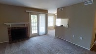 This single-floor residence offers a practical and comfortable layout designed to maximize both function and livability. The main living area is anchored by a brick-surround fireplace, providing a warm focal point for the room. Plush wall-to-wall carpeting covers the living space, adding a sense of comfort underfoot, while the neutral warm-toned walls lend the interior a clean, understated aesthetic. Natural light flows generously into the space through multiple windows fitted with horizontal blinds, as well as a full-length sliding glass door that opens onto an outdoor balcony, offering views of surrounding greenery. The open-concept design connects the living area to the kitchen via a pass-through counter with a breakfast bar overhang, creating a seamless flow between spaces while maintaining a sense of defined zones. The kitchen features a sink positioned at the pass-through, allowing for easy interaction with the living area. HVAC vents are visible in the ceiling, ensuring year-round climate control. The overall ambiance is bright and welcoming, with a straightforward, functional design that suits a variety of living arrangements.