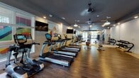 This fitness center offers a thoughtfully designed, single-floor workout space that balances functionality with a polished aesthetic. The main area is anchored by a row of True-brand treadmills and elliptical machines arranged along one wall, each equipped with individual display screens for an engaging cardio experience. Multiple wall-mounted flat-screen televisions are positioned throughout the space, ensuring visibility from virtually every piece of equipment.

The flooring throughout is rich hardwood, lending warmth and a clean, upscale feel to the environment. Dark-painted ceilings contrast with bright white walls, creating a modern, energetic atmosphere. Ceiling fans with pendant-style fixtures provide air circulation, while recessed lighting ensures the space remains well-lit. Large windows fitted with blinds allow natural light to filter in, and a set of glass-paned double doors at one end opens to an outdoor area, further enhancing the sense of openness.

A dedicated strength training section features a cable machine, a weight bench, and a selectorized resistance machine, offering a comprehensive range of workout options. Mirrored panels and interior windows between zones visually expand the space and allow sightlines across the full facility. Colorful artwork adds a vibrant, motivating touch.