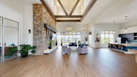 This single-floor communal facility is designed with a contemporary aesthetic that balances bold architectural elements with open, inviting spaces. The grand entrance features soaring ceilings, stacked-stone pillar accents, large glass doors, and wood-look flooring that flows seamlessly throughout the space.

The centerpiece of the main lounge is a floor-to-ceiling stacked-stone fireplace wall, complete with a mounted flat-screen television and a built-in hearth. An elegant coffered ceiling with dark wood beams frames the lounge area, while drum pendant lights and strategically placed accent lighting enhance the refined ambiance. Expansive windows and glass doors line the walls, flooding the interior with abundant natural light and offering views of the lush outdoor greenery.

The lounge is furnished with a comfortable seating arrangement, including plush armchairs and sofas accented with blue and teal throw pillows, gathered around a circular white coffee table — creating a warm and social gathering environment.

The kitchen area is thoughtfully appointed with dark cabinetry, granite countertops, a light blue subway tile backsplash, stainless steel appliances including a dishwasher and refrigerator, and a butcher-block island with wicker and metal bar seating, illuminated by globe pendant lights. The space also features a billiards table, visible in the background, adding a recreational dimension to this well-rounded communal facility.