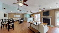 This single-floor community clubhouse offers an open, well-connected layout designed for gathering and relaxation. The entrance foyer greets visitors with vaulted ceilings, warm hardwood flooring, and tasteful accent seating, setting a welcoming tone throughout. Multiple ceiling fans are distributed across the space, ensuring comfortable airflow in every area.

The main living area is generously proportioned, centered around a classic fireplace framed by a white mantel and flanked by a wall-mounted flat-screen television. A set of French doors opens to an outdoor view of what appears to be a pool area, allowing abundant natural light to fill the room. Comfortable seating, including a cream sofa and teal accent chairs, completes the lounge atmosphere.

Adjacent to the living area, a dedicated dining zone features a round glass-top table with dark-framed chairs, alongside a pass-through window offering a view into a secondary dining or activity room. The kitchen is defined by a raised granite-topped breakfast bar, wood cabinetry, stainless steel appliances, and recessed lighting, providing a functional service area. The open-plan design ensures seamless flow between the kitchen, dining, and lounge zones, making this clubhouse an ideal communal hub.