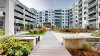 Located at 652 Angier Ave NE in Atlanta's Old Fourth Ward, this contemporary multi-story residential community is designed around a thoughtfully landscaped outdoor amenity level. The grounds are anchored by a large resort-style swimming pool enclosed within the building's courtyard, offering a serene retreat with views of the surrounding architecture's clean white façade, teal-accented windows, and private balconies.

Meandering concrete walkways connect a series of distinct outdoor gathering spaces, each with its own character. Carefully designed planting beds feature ornamental trees, seasonal shrubs, river rock arrangements, and mulched borders that lend a natural, polished aesthetic throughout. A covered lounge area with warm wood-beam ceilings, integrated greenery, and ceiling fans provides a comfortable semi-outdoor retreat, while an open-air deck with hardwood flooring and upholstered seating offers additional relaxation space.

Multiple social zones include an outdoor dining area with a fireplace, a fire pit lounge framed by evergreens and boulders, and a shaded patio with bistro tables bordered by a warm-toned wood fence. The overall ambiance blends modern urban architecture with inviting, resort-inspired outdoor living, making this Atlanta community a well-appointed destination for contemporary residential life.